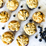 10 blueberry crumble muffins on a marble backdrop with a pile of blueberries to the bottom right.
