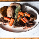 A Beef Chuck Roast on an oval white platter with carrots and potatoes on top