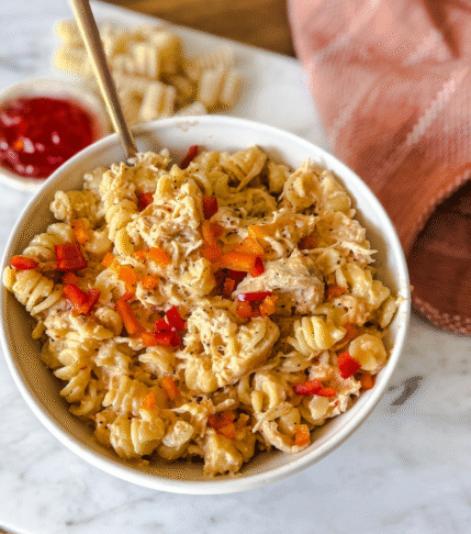 Jalapeno Pepper Jelly Pasta in a white bowl with a marble backdrop. An orange fabric napkin is placed above and jalapeno pepper jelly and pasta to the left.