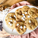 A pile of S' More Cookies on a white plate with marshmallows, chocolate chips and graham crackers crumbled