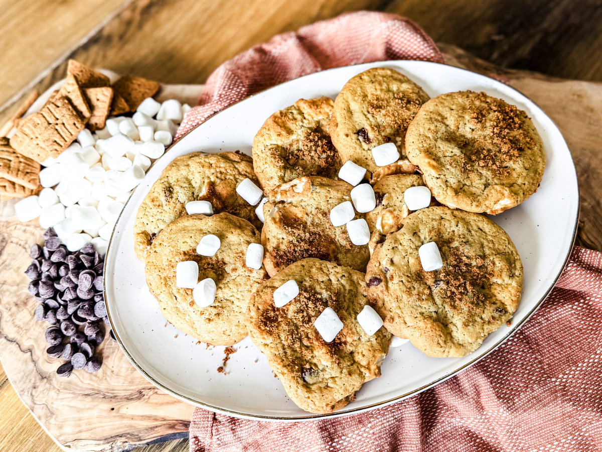 A pile of S' More Cookies on a white plate with marshmallows, chocolate chips and graham crackers crumbled