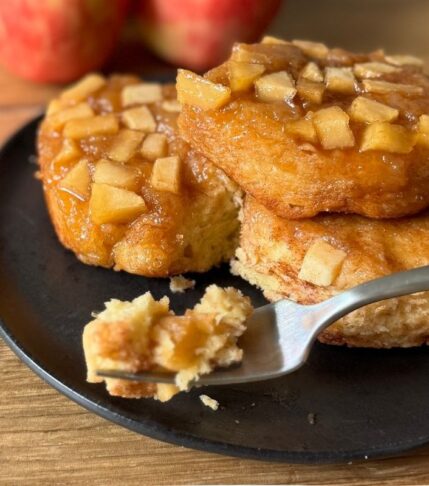 3 Einkorn Flour Apple Sticky Buns on a Round black plate with a wooden background. A fork hold up a bite of the sticky bun.
