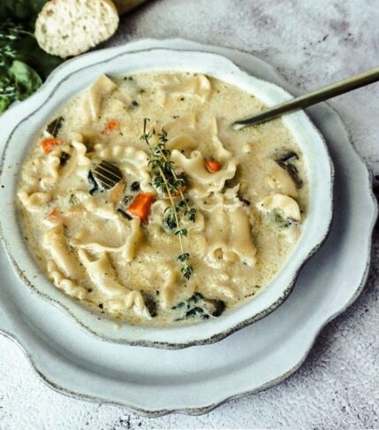 Chicken Lasagna Soup in a scalloped white bowl on top of a scalloped dish with bread to the top left and a gold thing handle spoon in the soup. Thyme is placed on top of the soup.