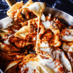 A pan of Skillet shells and cheese with a marble background. A wooden spoon pulls a scoop of the shells up. Letting the gooey cheese drizzle down.