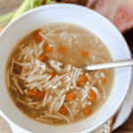Chicken Noodle Soup in a white bowl with a wooden background. A gold spoon is in the bowl and carrots and celery are in the background.