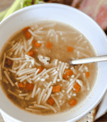 Chicken Noodle Soup in a white bowl with a wooden background. A gold spoon is in the bowl and carrots and celery are in the background.
