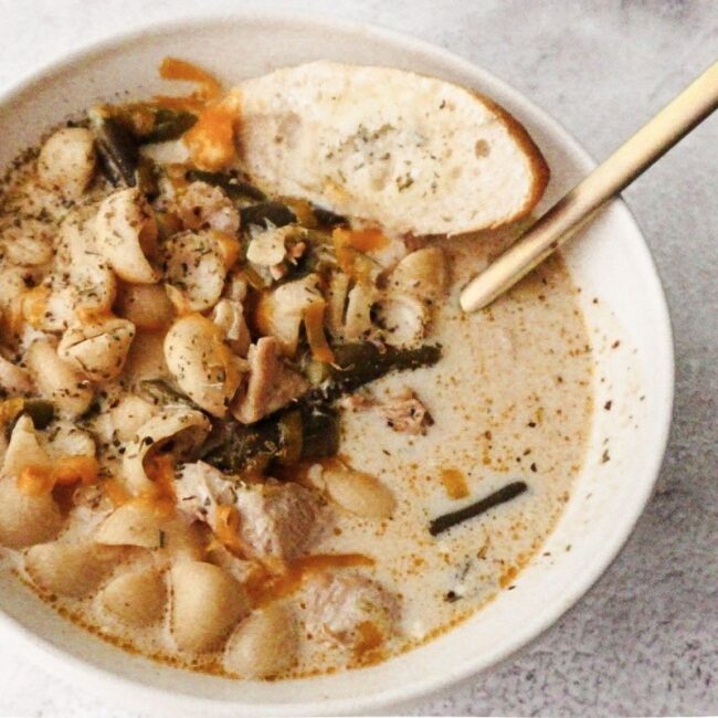 A bowl of No-Bean White Chicken Chili with a gold spoon to the top right and a slice of bread to the top left.