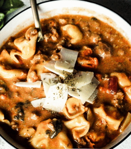 Lasagna soup in a white bowl with cheese and herbs sprinkled on top. A gold spoon is placed to the top left, a white backdrop and a piece of bread to the left.