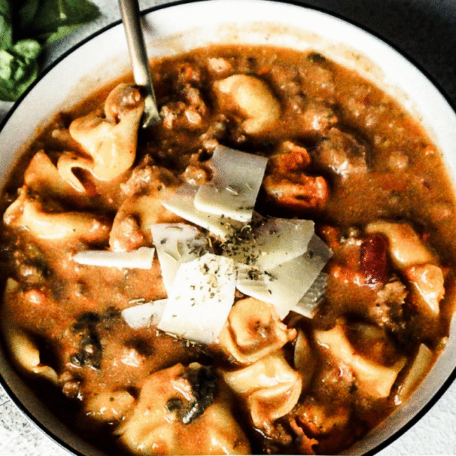 Lasagna soup in a white bowl with cheese and herbs sprinkled on top. A gold spoon is placed to the top left, a white backdrop and a piece of bread to the left.