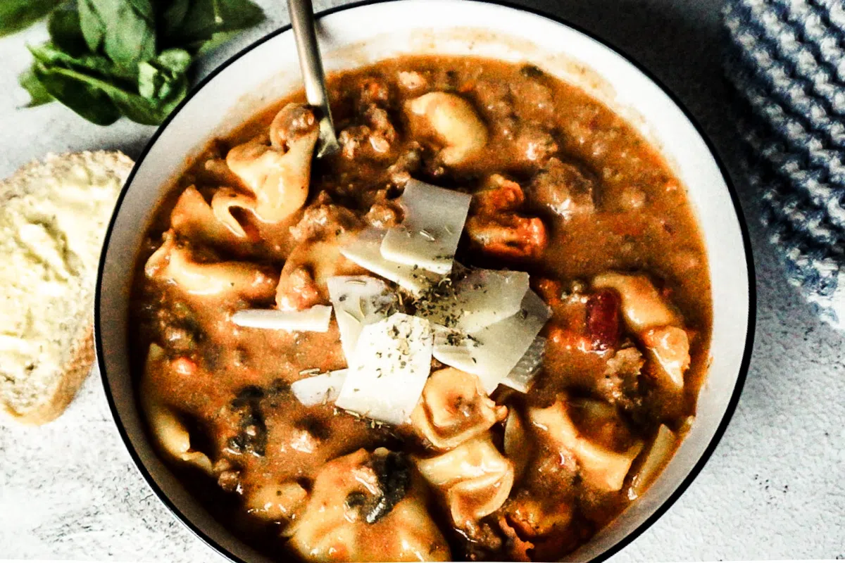 Lasagna soup in a white bowl with cheese and herbs sprinkled on top. A gold spoon is placed to the top left, a white backdrop and a piece of bread to the left.