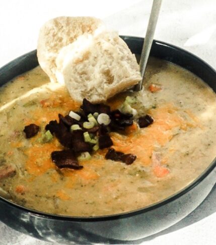 Loaded baked potato soup in a black bowl with a white back drop. A gold spoon is placed in and tilted to the side. It is piled high with cheese, bacon and green onions and a crusty piece of bread is to the side.