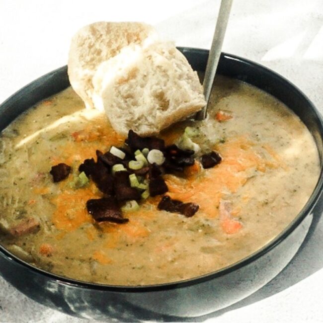 Loaded baked potato soup in a black bowl with a white back drop. A gold spoon is placed in and tilted to the side. It is piled high with cheese, bacon and green onions and a crusty piece of bread is to the side.