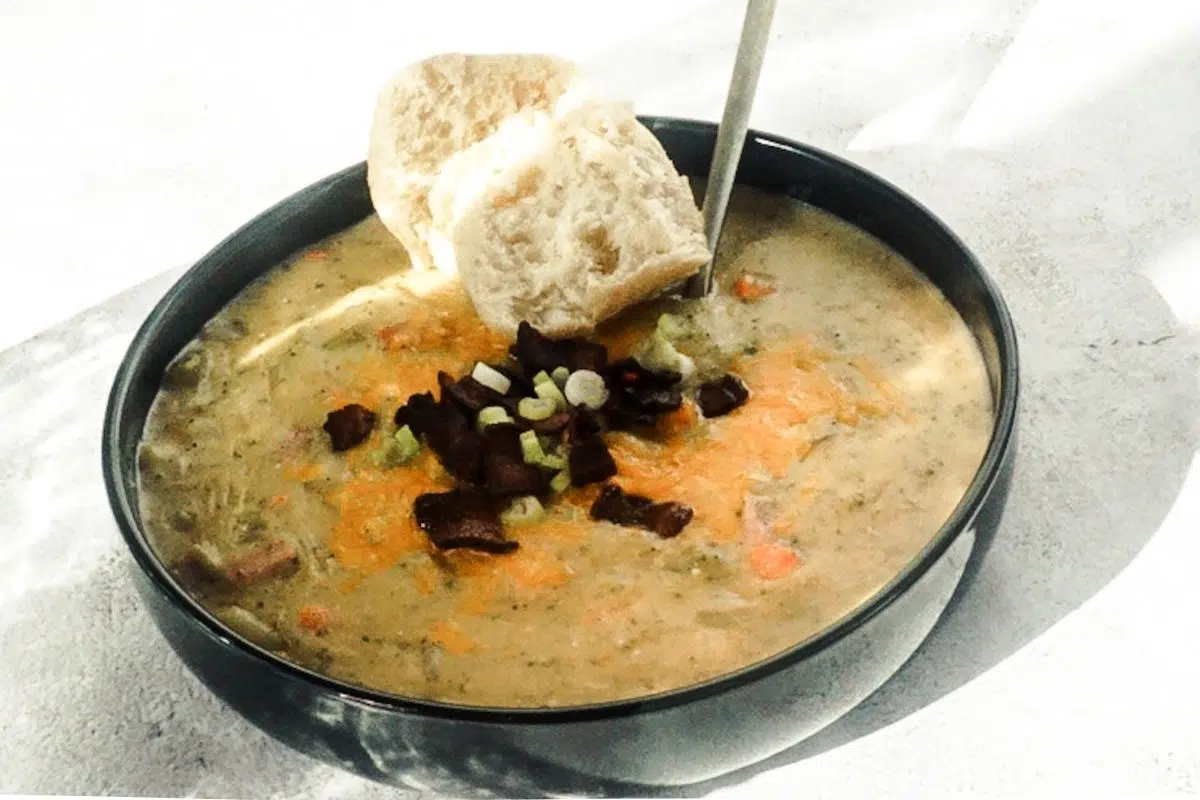Loaded baked potato soup in a black bowl with a white back drop. A gold spoon is placed in and tilted to the side. It is piled high with cheese, bacon and green onions and a crusty piece of bread is to the side.