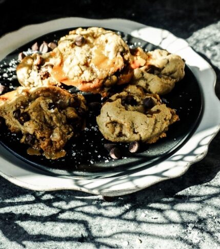Sea Salt Caramel Chocolate Chip Cookies on a black plate, then placed on a scalloped white plate. A black background with shadows of branches cover the image.