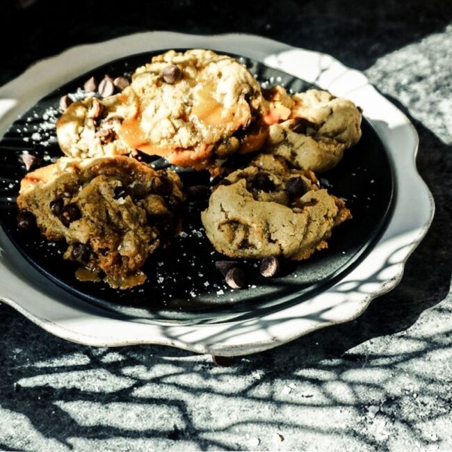Sea Salt Caramel Chocolate Chip Cookies on a black plate, then placed on a scalloped white plate. A black background with shadows of branches cover the image.
