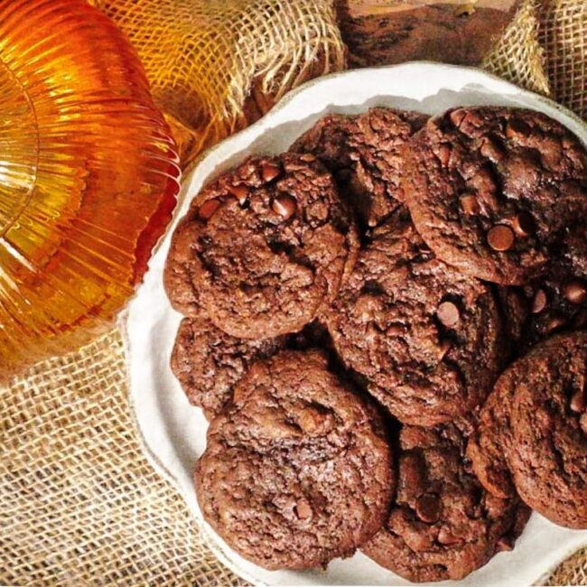 Asher's Double Chocolate Pumpkin Cookies piled on a white plate with a pumpkin to the left. A brown and black backdrop.
