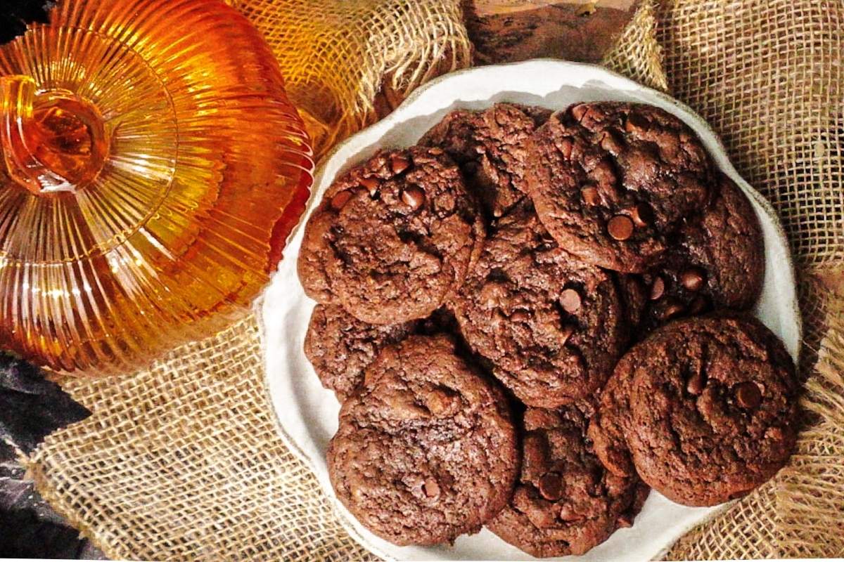 Asher's Double Chocolate Pumpkin Cookies piled on a white plate with a pumpkin to the left. A brown and black backdrop.