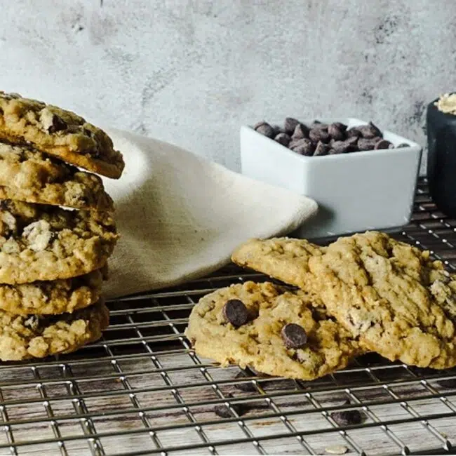 oatmeal chocolate chip cookies stacked on a wire rack. A pole of chocolate chips and oats are in the backdrop.