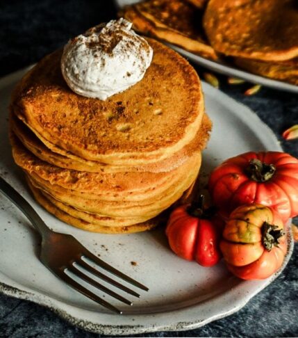 Pumpkin Pancakes stacked on an off white scalloped plate with a dollop of cinnamon whipped cream on top. A for sits to the left, 3 small pumpkins on the plate and a plate of pancakes on the distance.