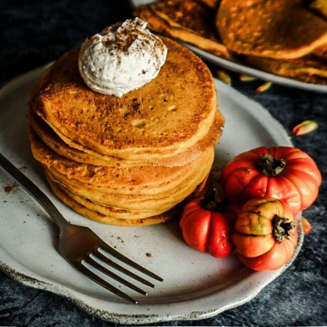 Pumpkin Pancakes stacked on an off white scalloped plate with a dollop of cinnamon whipped cream on top. A for sits to the left, 3 small pumpkins on the plate and a plate of pancakes on the distance.