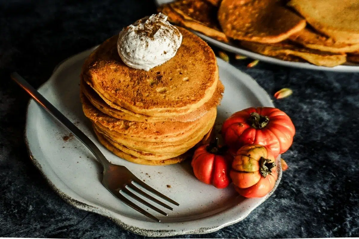 Pumpkin Pancakes stacked on an off white scalloped plate with a dollop of cinnamon whipped cream on top. A for sits to the left, 3 small pumpkins on the plate and a plate of pancakes on the distance.