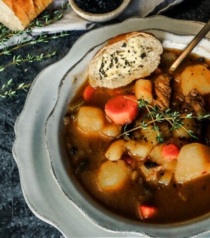 Pumpkin Stew in a bowl with a scalloped dish underneath. A piece of bread is resting to the left with a gold spoon.