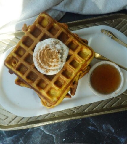 Pumpkin waffles on a white backdrop, with a gold plate behind. A small bowl of syrup and gold tongs