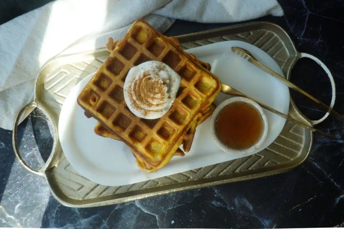 Pumpkin waffles on a white backdrop, with a gold plate behind. A small bowl of syrup and gold tongs