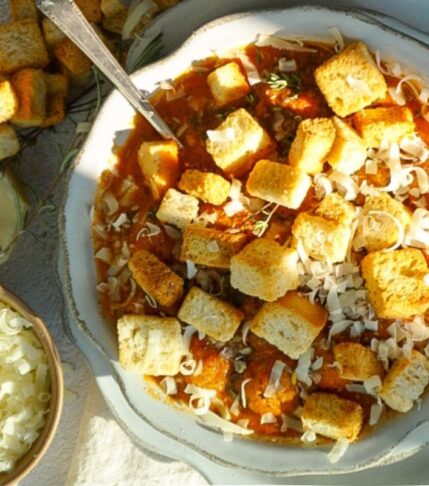 Chicken Parm Soup in a scalloped bowl with parmesan cheese in a small bowl to the left. Croutons are scattered on the top and it is sprinkled with parmesan cheese.