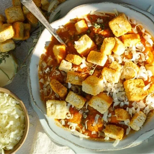 Chicken Parm Soup in a scalloped bowl with parmesan cheese in a small bowl to the left. Croutons are scattered on the top and it is sprinkled with parmesan cheese.
