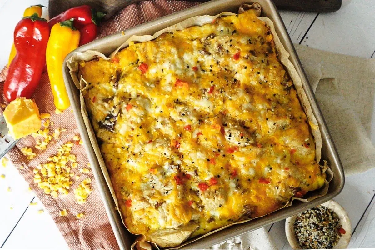 Sausage Breakfast bake in a square baking dish with a white back drop and dish towel behind. Cheese grater to the left with peppers.