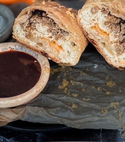 a sausage and boursin breakfast burrito split in 2. A side of hot sauce in a small dish is to the left. There is parchment paper and sour cream on the side.
