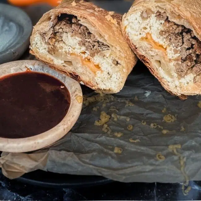 a sausage and boursin breakfast burrito split in 2. A side of hot sauce in a small dish is to the left. There is parchment paper and sour cream on the side.