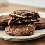 Double Chocolate Cheesecake Cookies on a scalloped white plate with a wooden backdrop. Piled with a cookie split open so the cheesecake filling shows.