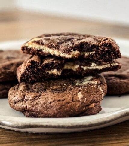 Double Chocolate Cheesecake Cookies on a scalloped white plate with a wooden backdrop. Piled with a cookie split open so the cheesecake filling shows.