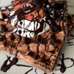 Chocolate waffles on a scalloped plate drizzled in chocolate and topped with whipped cream and strawberries.
