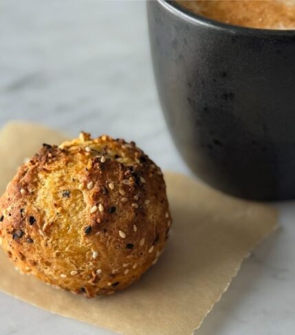 Everything bagel bomb filled with cream cheese on a piece of parchment paper with a coffee mug in the background.