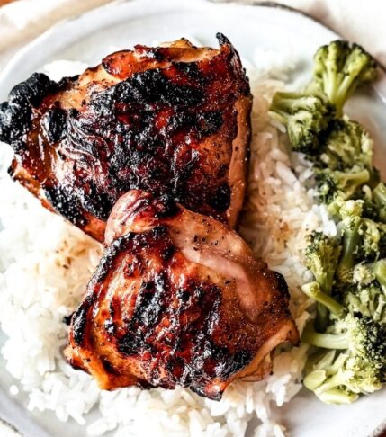 2 chicken thighs plated on rice with a side of broccoli. A white scalloped plate is in the backdrop.