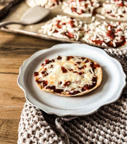 Tortilla pizza on a scalloped off white plate with pizzas on a sheetpan behind. A tan towl is behind the plate.