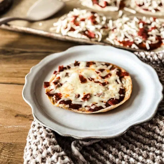 Tortilla pizza on a scalloped off white plate with pizzas on a sheetpan behind. A tan towl is behind the plate.